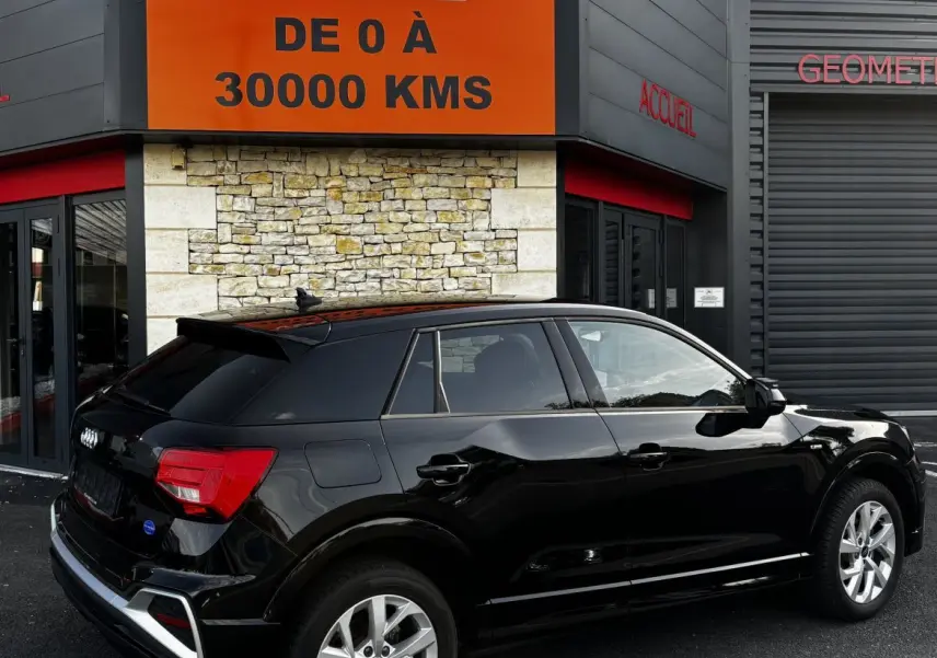 Audi Q2 noir vue 3/4 arrière droit, stationné devant un bâtiment avec panneau orange et pierre apparente.