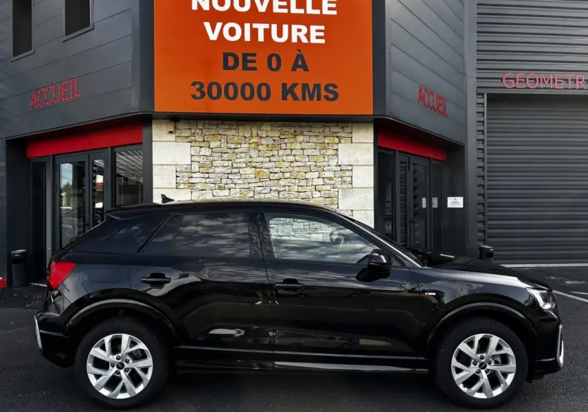 Audi Q2 35 TFSI S Line noir vue de profil côté gauche, stationné devant un bâtiment avec panneau orange.