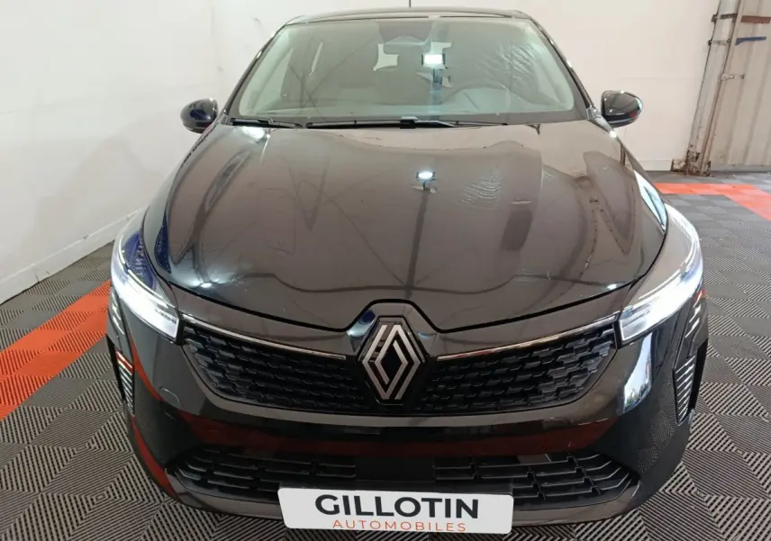 Vue avant d'une Renault Clio noire 2025 avec calandre moderne et phares LED allumés en intérieur showroom.