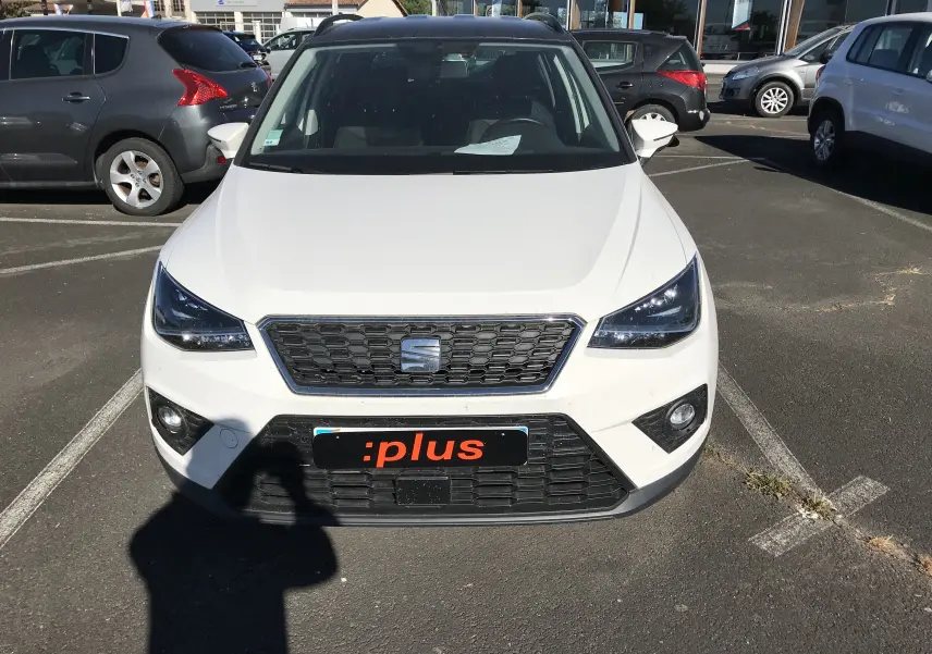 Vue frontale d'une SEAT Arona blanche avec toit noir, sur un parking, affichant la calandre et les phares LED.
