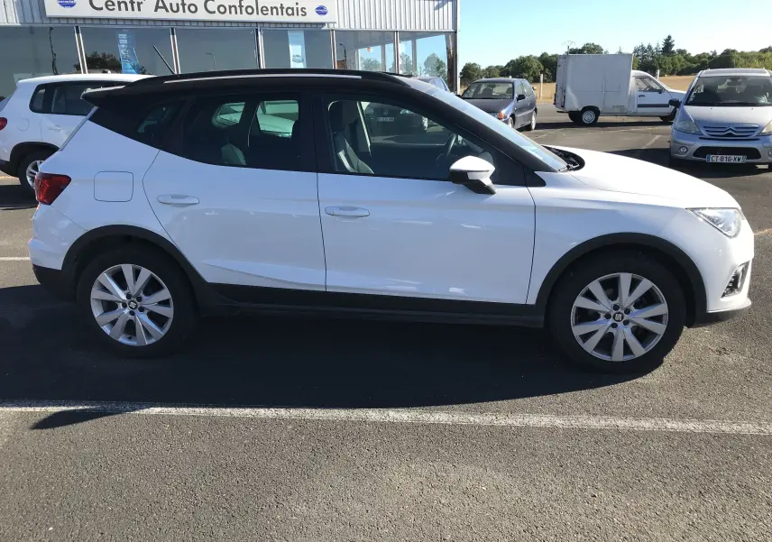 SEAT Arona blanc avec toit noir vu de profil côté gauche, garé devant un centre auto en plein jour.