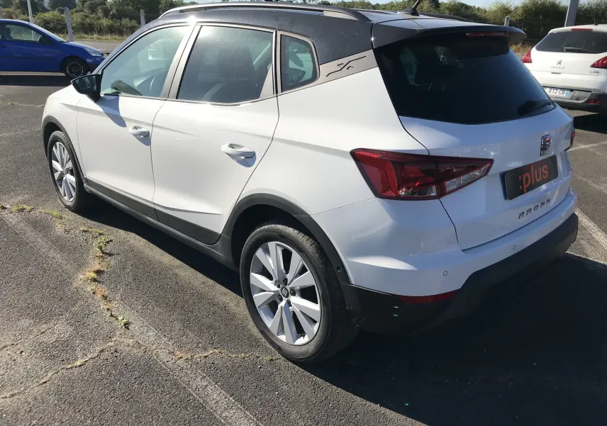 Vue 3/4 arrière droite d'un SEAT Arona blanc avec toit noir et jantes alliage sur parking ensoleillé.