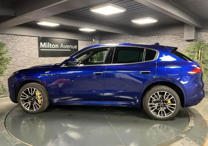 Profil droit du Maserati Grecale bleu avec jantes noires et étriers de freins jaunes visibles en intérieur showroom.