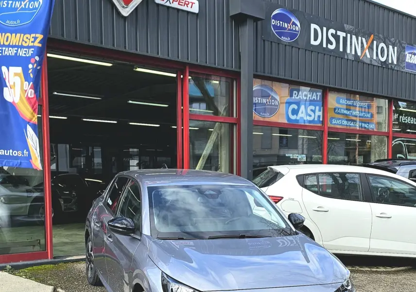 Peugeot 208 110 hybrid gris vue 3/4 avant droit, avec calandre noire et feux LED distinctifs, devant un garage.