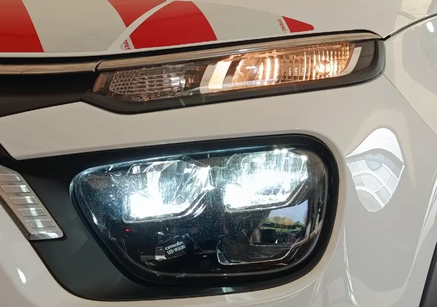 Gros plan sur le phare avant droit allumé d'une Citroën C3 blanche, mettant en valeur les LED et le clignotant orange.