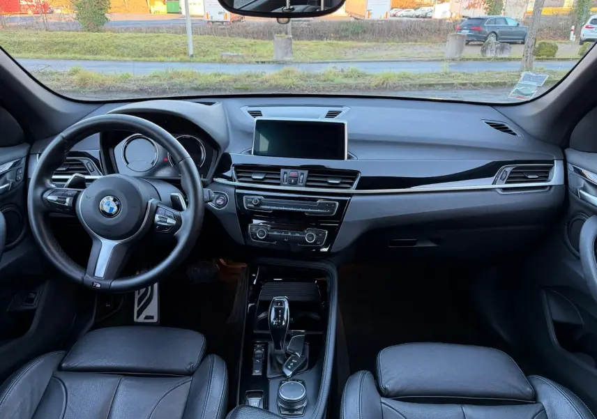 Vue intérieure frontale du tableau de bord et volant noir cuir du BMW X1 18D M Sport, avec écran central et console brillante.