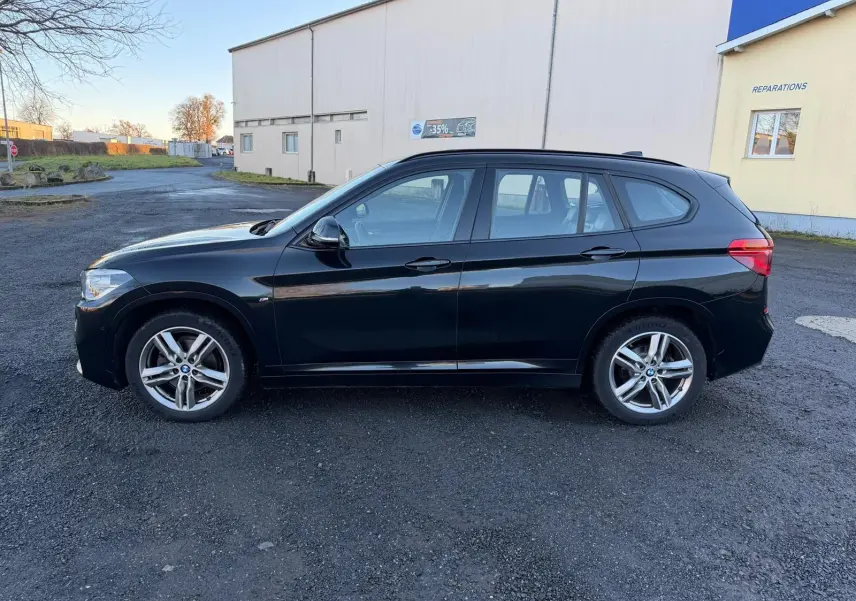 BMW X1 18D M Sport noir métal vue de profil côté gauche sur un sol goudronné devant un bâtiment clair.