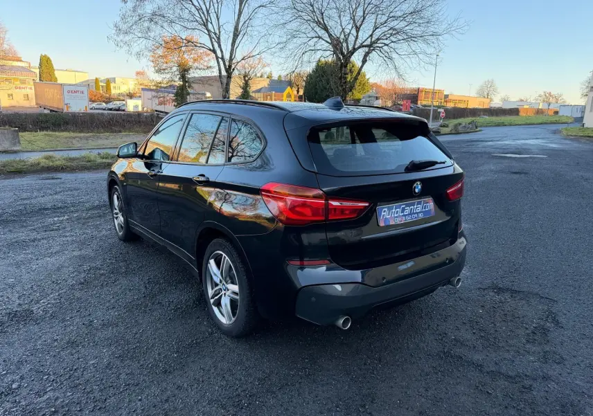 BMW X1 noir métal vue 3/4 arrière droit sur parking, avec feux arrière allumés et jantes alliage 18 pouces.