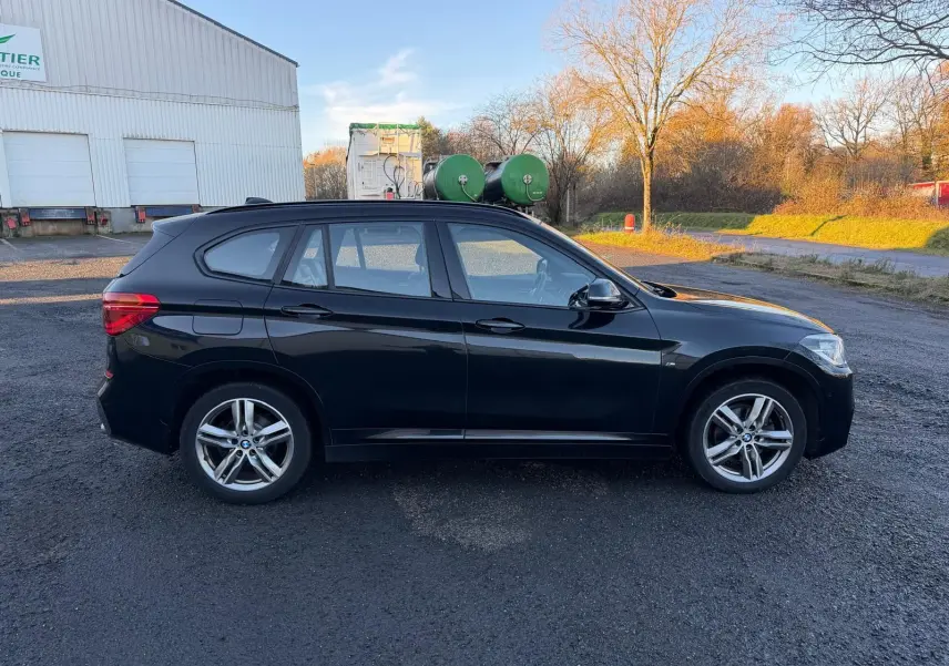BMW X1 18D M Sport noir métal vue de profil côté gauche sur un sol goudronné en extérieur au coucher du soleil.