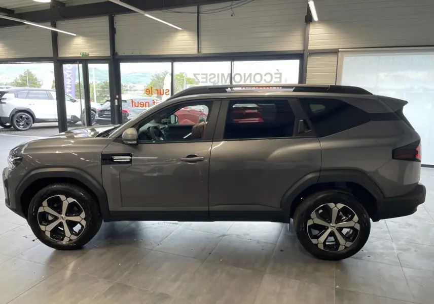 Vue latérale droite du Dacia Bigster gris schiste 2026 avec jantes alliage 18 pouces et barres de toit noires.