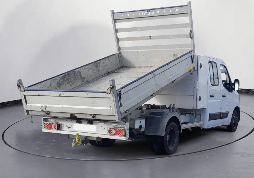 Renault Master blanc double cabine vu 3/4 arrière avec benne basculante relevée et attelage visible.
