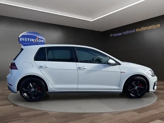 Vue de profil droit d’une Volkswagen Golf GTI Performance blanche avec jantes noires et étriers rouges, en intérieur showroom.