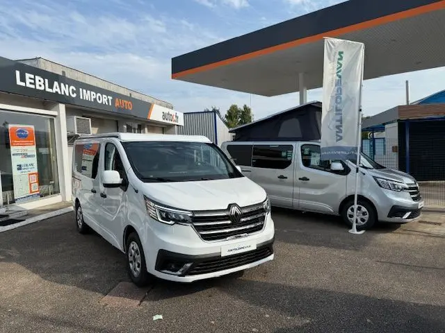 Renault Trafic Fourgon blanc glacier en 3/4 avant gauche, stationné devant un garage avec un autre van en arrière-plan.