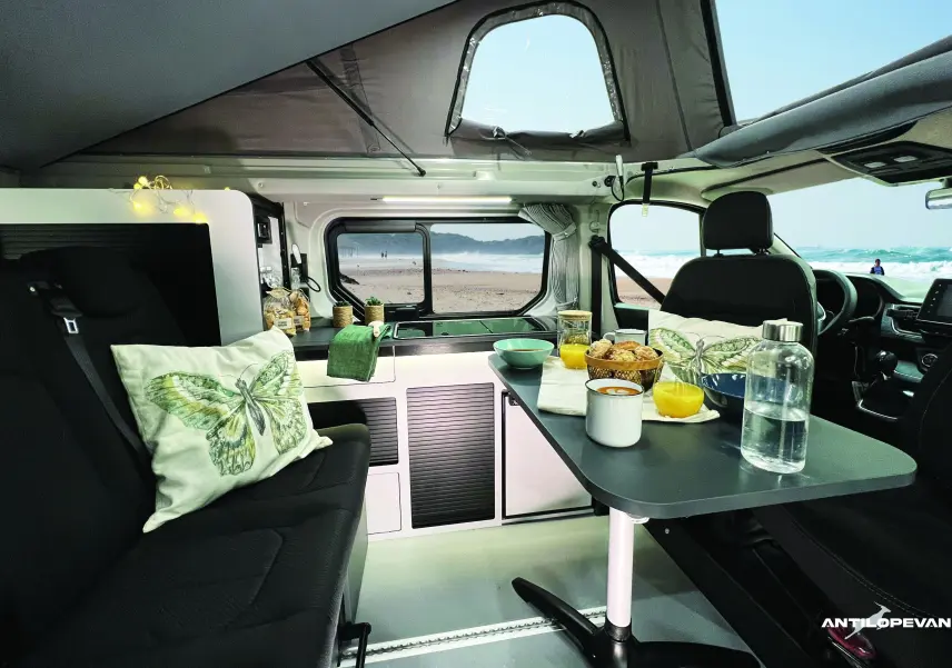 Intérieur du Renault Trafic Fourgon blanc glacier aménagé Antilope Van, vue latérale sur la cuisine et coin repas avec table et sièges noirs.