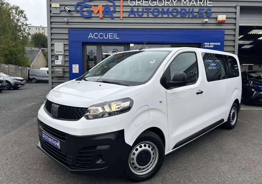 Fiat Scudo Combi blanc en 3/4 avant droit, avec calandre noire et jantes acier, devant un garage automobile.