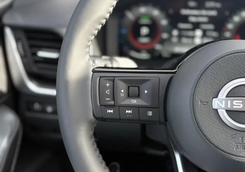 Gros plan sur les commandes au volant à gauche du Nissan Qashqai blanc nacré, avec tableau de bord numérique flou en arrière-plan.