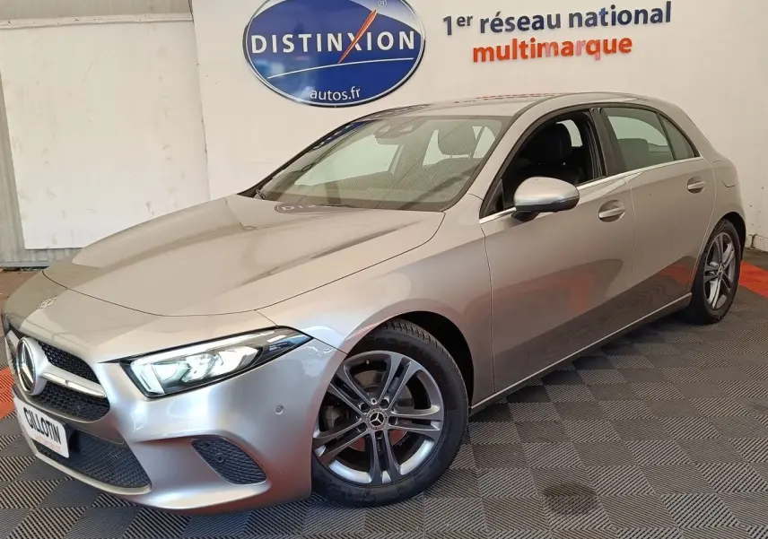 Vue 3/4 avant d'une Mercedes Classe A 180 Business Line gris clair avec jantes alliage et phares LED allumés.
