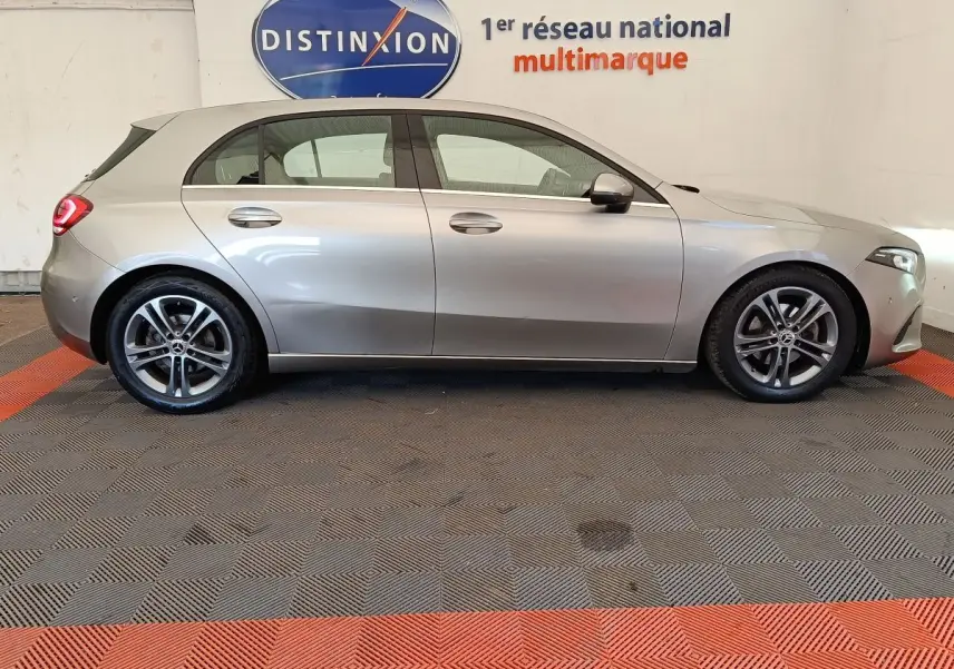 Vue de profil côté gauche d'une Mercedes Classe A 180 Business Line gris clair stationnée en intérieur sur sol à damiers.