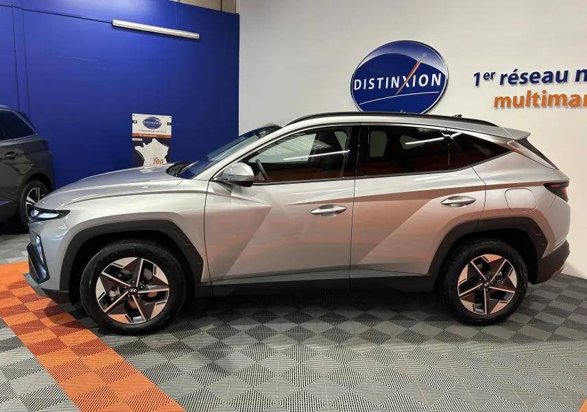 Vue de profil côté gauche du Hyundai Tucson 2025 hybride en Shimmering Silver avec jantes 18 pouces et vitres surteintées.