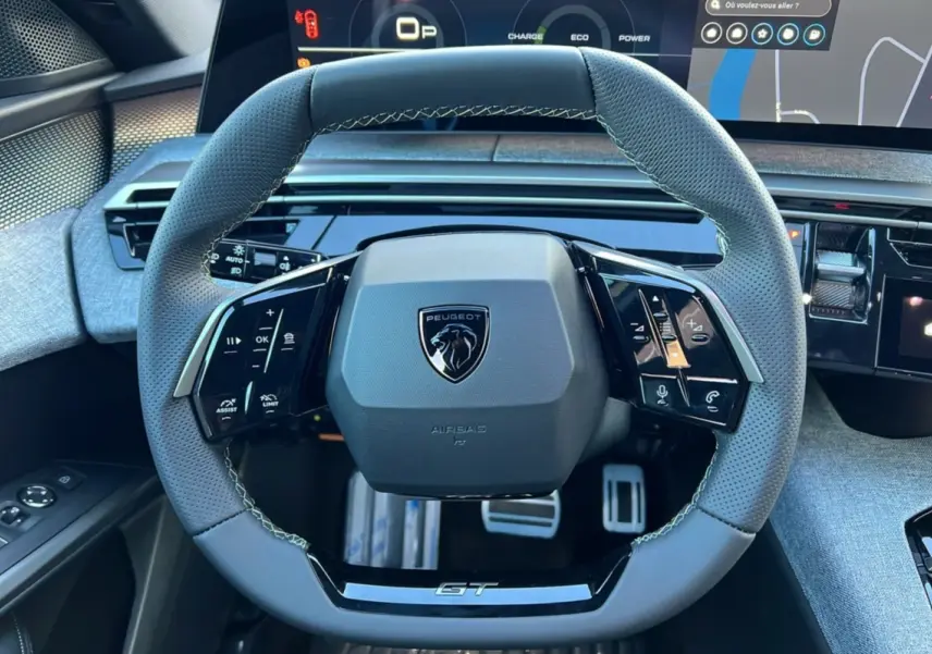 Vue rapprochée du volant cuir perforé gris avec logo Peugeot et inscription GT, tableau de bord numérique en arrière-plan.