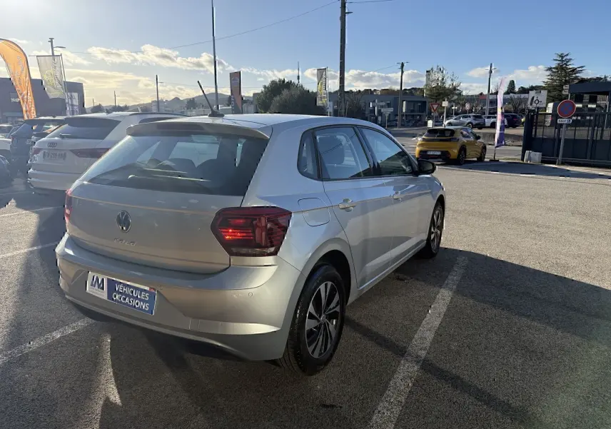 Volkswagen Polo gris métal vue 3/4 arrière droit, feux arrière allumés, stationnée en extérieur au soleil.