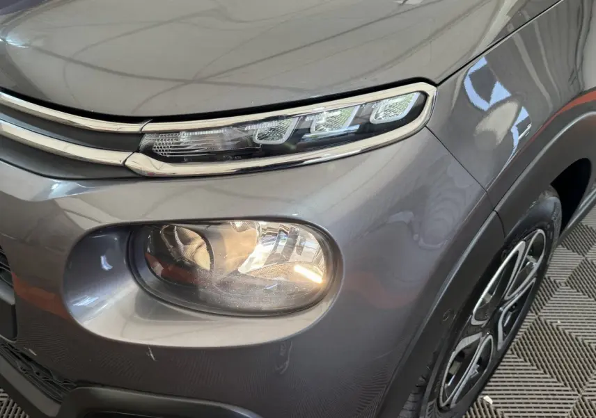 Gros plan sur le phare avant droit et la calandre chromée d'une Citroën C3 gris foncé, avec jante visible.