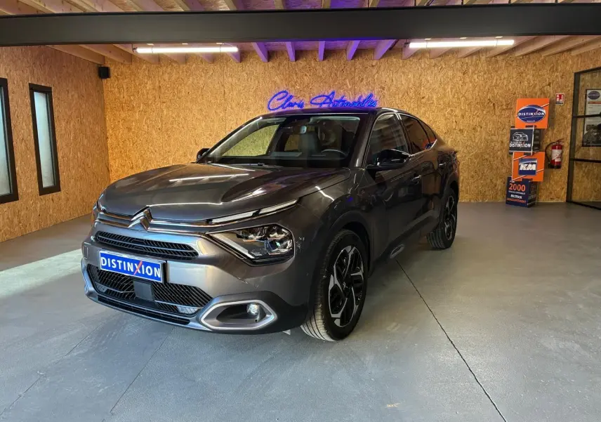 Vue 3/4 avant droite d'une Citroën C4 X gris métallisé 2024 avec phares LED et jantes noires en intérieur showroom.