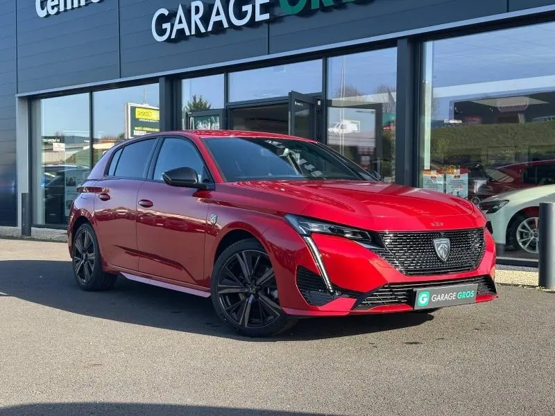 Peugeot 308 rouge Elixir vue 3/4 avant droit, avec calandre noire et jantes alliage noires devant un garage.