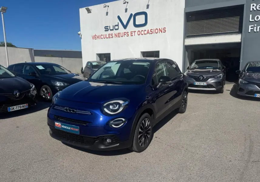 Vue 3/4 avant d'une FIAT 500X bleu métallisé garée devant un concessionnaire, avec jantes classiques noires.