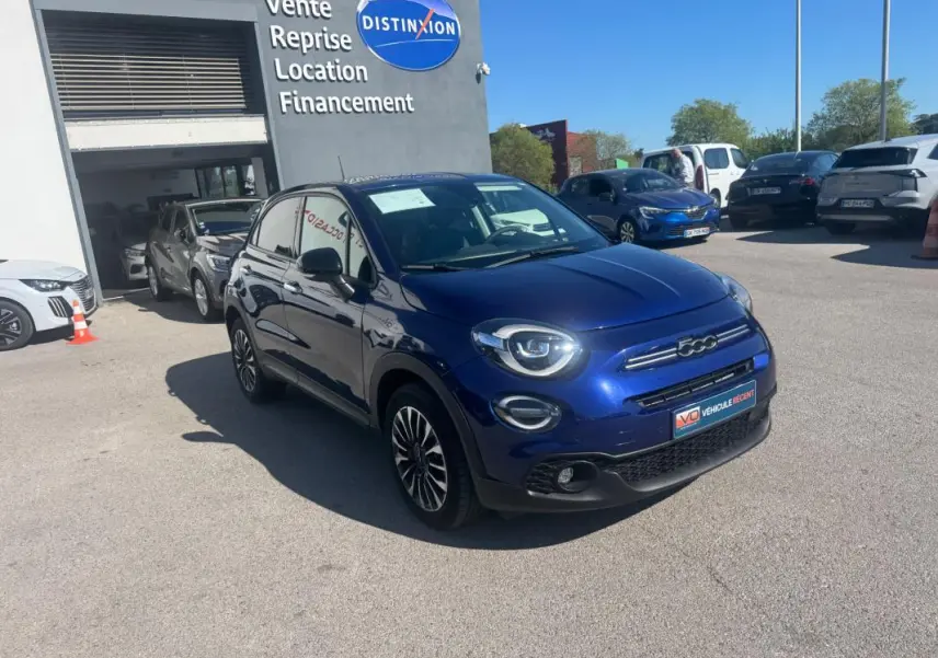 Vue 3/4 avant droit d'une FIAT 500X bleu métallisé avec jantes classiques 16 pouces, stationnée devant un garage.