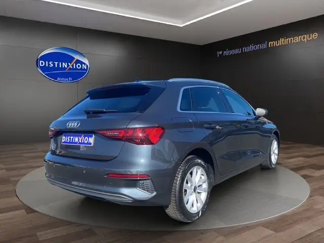 Vue 3/4 arrière droite d'une Audi A3 Sportback gris Manhattan avec vitres arrière surteintées en intérieur showroom.