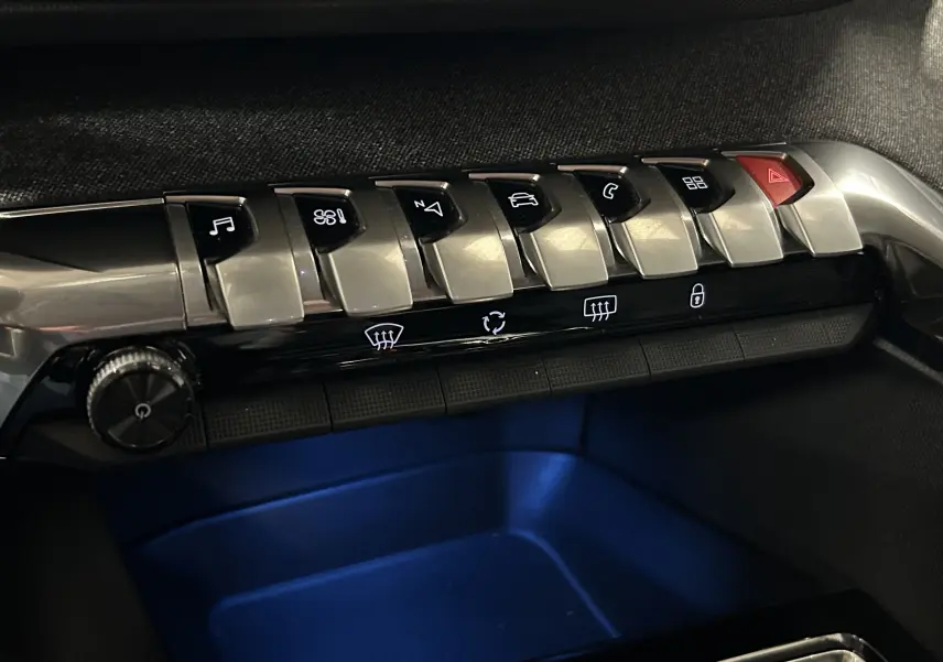 Gros plan sur les commandes centrales en métal brossé du tableau de bord du Peugeot 3008, éclairage bleu dessous.