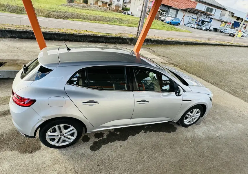 Vue de côté droit en léger plongé d'une Renault Mégane IV berline gris métallisé stationnée sous abri avec jantes alliage.