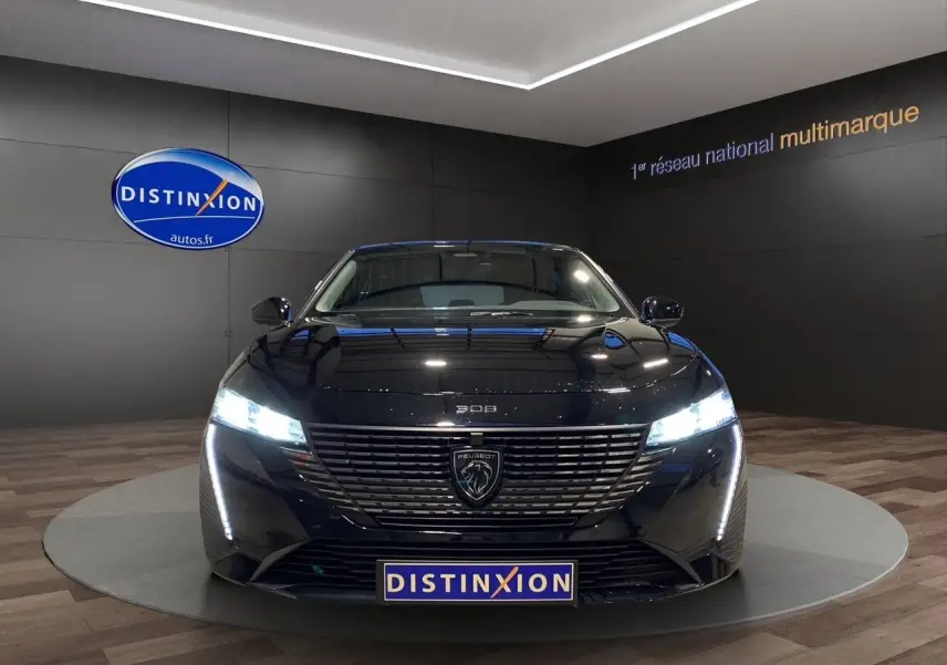 Peugeot 308 1.5 BlueHDi 130 Allure en noir Perla Nera, vue de face avec phares LED allumés dans showroom.
