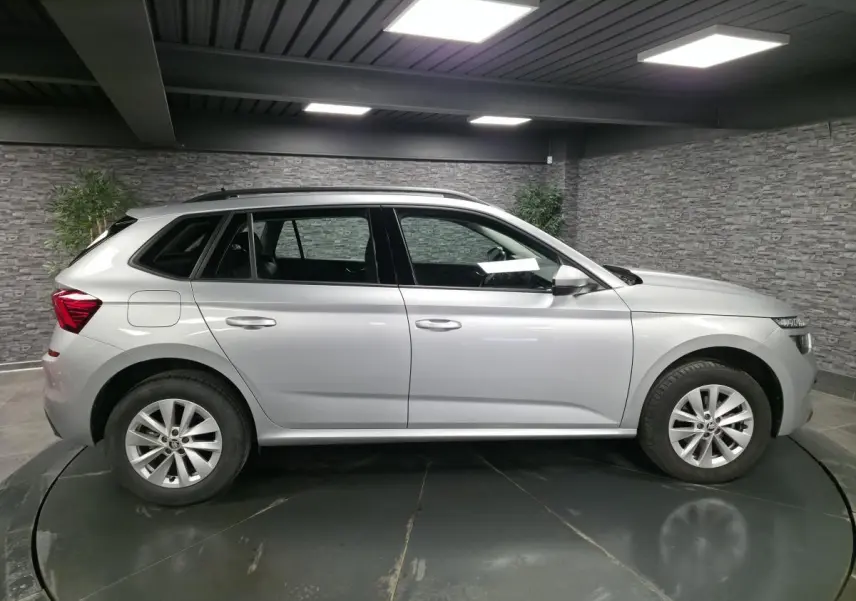Vue de profil côté gauche d'un Skoda Kamiq gris métallisé 2023 avec jantes alliage et barres de toit noires.