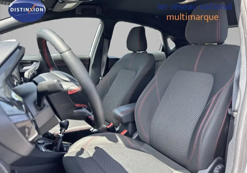 Intérieur avant du Ford Puma blanc 2022, sièges noirs avec surpiqûres rouges et volant cuir perforé avec coutures rouges.