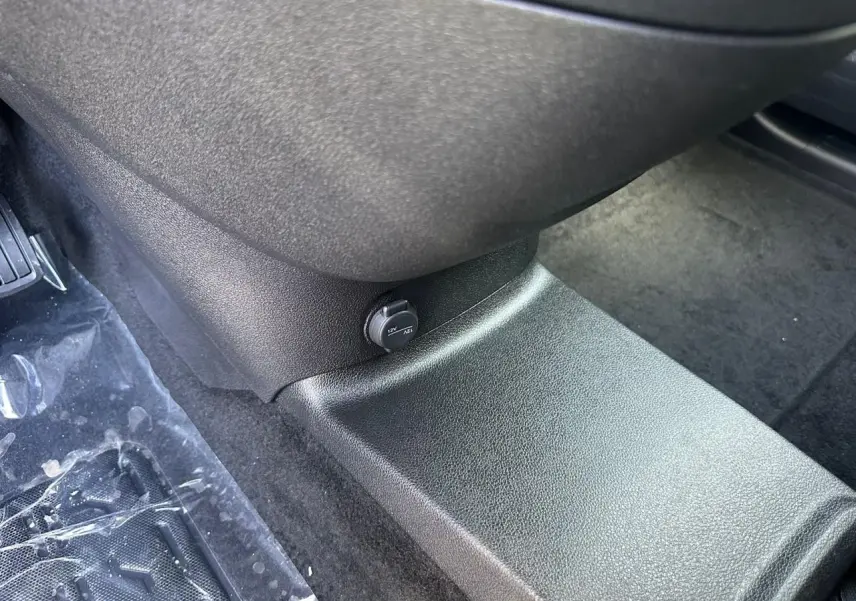 Prise 12V noire sur console centrale côté passager du Citroën Berlingo III blanc, plancher moquette visible.