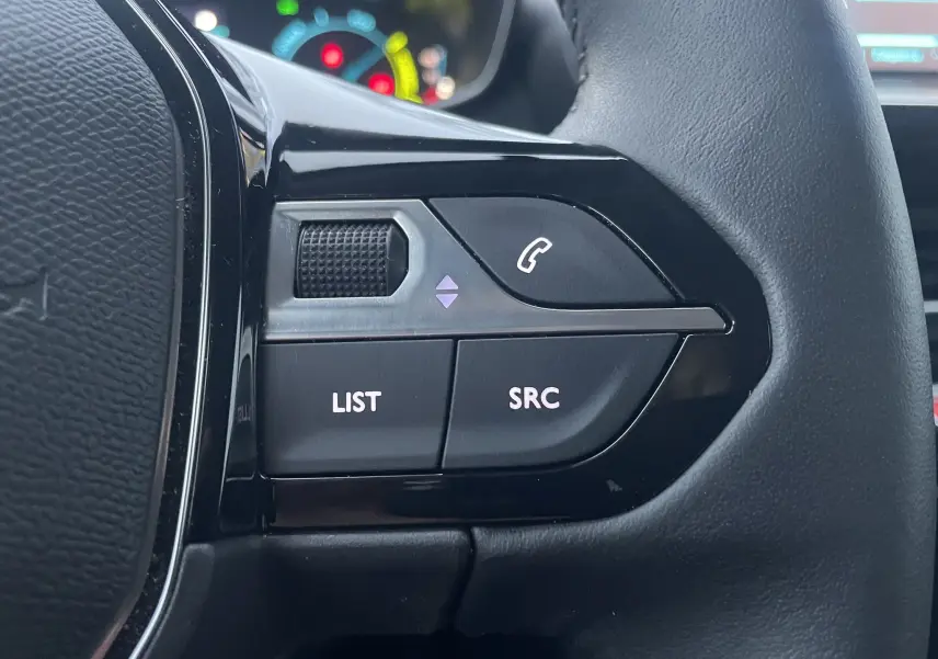 Gros plan sur les commandes gauche du volant noir du Peugeot 208 II électrique 2023, avec boutons LIST et SRC visibles.