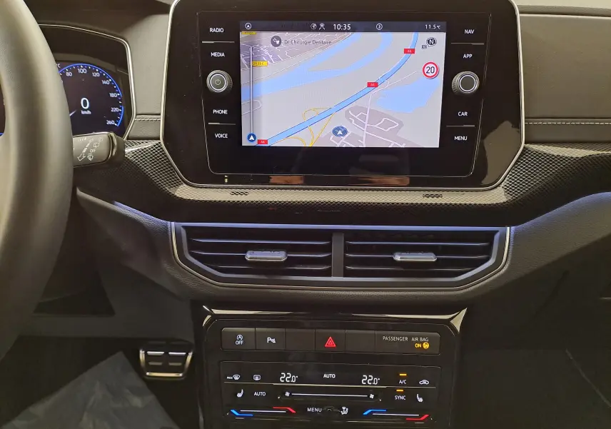 Vue intérieure du tableau de bord du Volkswagen T-Cross blanc, avec écran tactile central affichant la navigation et commandes climatisation.