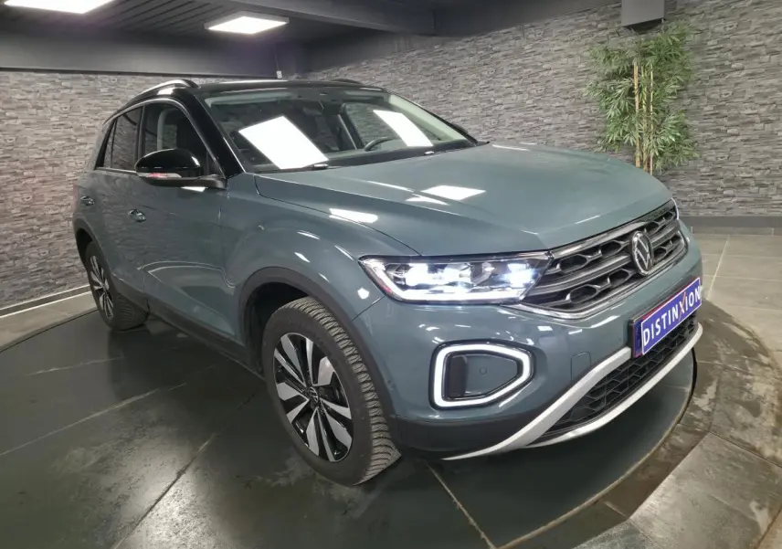 Volkswagen T-Roc bleu 3/4 avant droit avec feux LED allumés et toit noir dans un showroom.