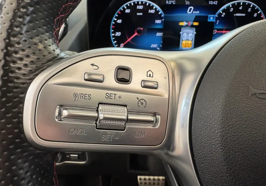 Gros plan sur les commandes argentées du volant cuir perforé noir d’un Mercedes GLA 250 e AMG, tableau de bord numérique visible.