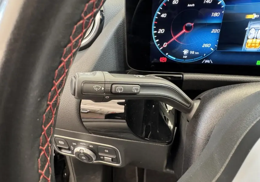 Gros plan sur le volant noir avec surpiqûres rouges et la commande d’essuie-glace, tableau de bord numérique visible en arrière-plan.