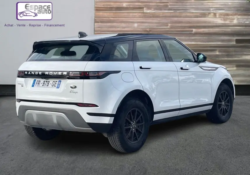 Vue 3/4 arrière droite d'un Range Rover Evoque blanc 2020 avec toit noir et jantes foncées, stationné devant un mur en bois.
