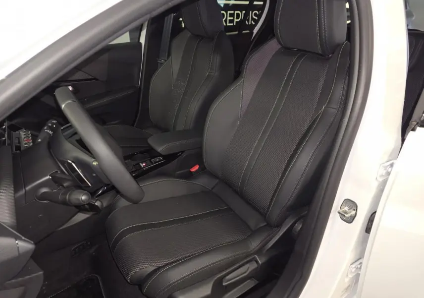 Vue intérieure côté conducteur d’une Peugeot 208 blanche 2025, siège tissu noir avec surpiqûres vertes et volant compact cuir.
