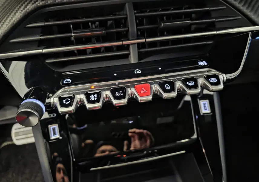 Gros plan sur la console centrale noire brillante du Peugeot 208 1.2i Hybrid 2025 avec boutons métalliques et éclairage LED.