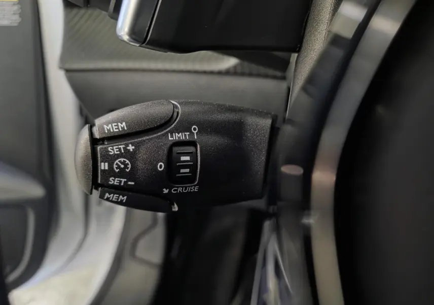 Gros plan sur la commande de régulateur de vitesse noire à gauche du volant dans l’habitacle du Peugeot 208 blanc Banquise.