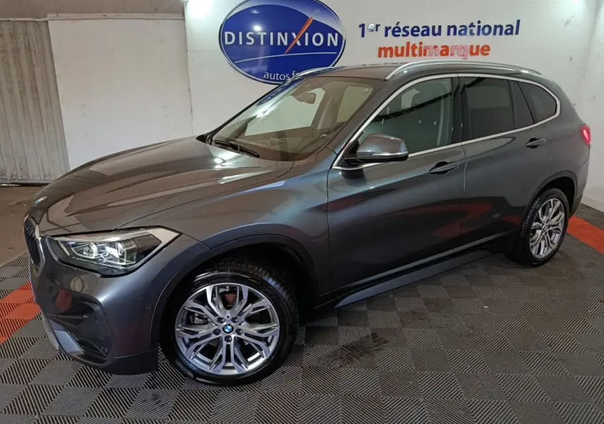 BMW X1 sDrive18i gris foncé vue 3/4 avant droit, feux allumés, roues alliage et rails de toit noirs visibles