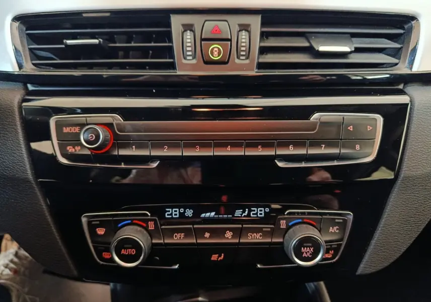 Vue rapprochée du tableau de bord noir brillant de la BMW X1 sDrive18i Lounge avec commandes climatisation digitale à 28°C.