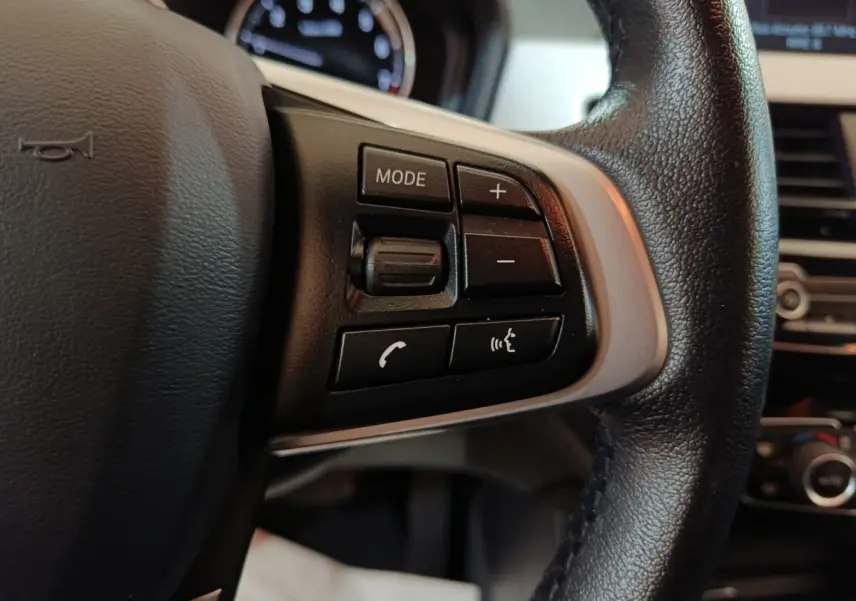 Gros plan sur les commandes gauche du volant cuir noir du BMW X1 sDrive18i Lounge 2021, avec tableau de bord flou en arrière-plan.