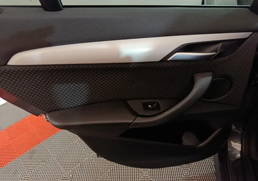 Vue intérieure du panneau de porte côté gauche d'une BMW X1 sDrive18i Lounge gris foncé, avec poignée et bouton de verrouillage.
